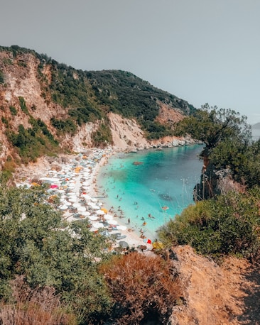 A scenic coastal area featuring a secluded beach with numerous white parasols and sunbathers enjoying the turquoise waters. The backdrop consists of rugged cliffs and lush greenery, providing a dramatic and natural setting.