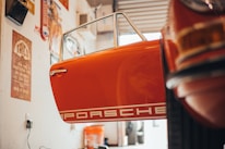 The scene depicts the interior of a garage or workshop with a vintage car door that is prominently visible. The door is orange and features the word 'Porsche' in white bold letters. Various tools and automotive accessories are scattered around, and there are vintage posters and signs on the walls, contributing to a retro ambiance. Warm lighting enhances the nostalgic feel of the space.