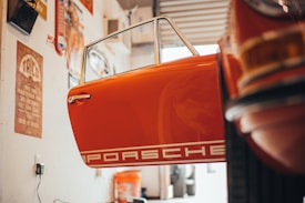 The scene depicts the interior of a garage or workshop with a vintage car door that is prominently visible. The door is orange and features the word 'Porsche' in white bold letters. Various tools and automotive accessories are scattered around, and there are vintage posters and signs on the walls, contributing to a retro ambiance. Warm lighting enhances the nostalgic feel of the space.