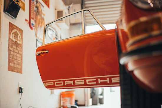 The scene depicts the interior of a garage or workshop with a vintage car door that is prominently visible. The door is orange and features the word 'Porsche' in white bold letters. Various tools and automotive accessories are scattered around, and there are vintage posters and signs on the walls, contributing to a retro ambiance. Warm lighting enhances the nostalgic feel of the space.