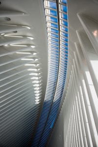 An interior architectural scene featuring a curved ceiling with a long skylight allowing natural light to illuminate the space. The structure is composed of repetitive, vertical white beams which create a sense of depth and dynamism. The skylight reveals a patch of blue sky, contrasting with the white interior.