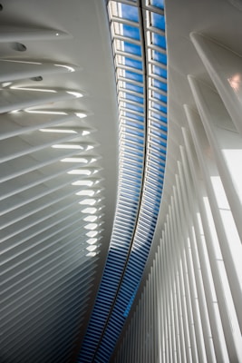 An interior architectural scene featuring a curved ceiling with a long skylight allowing natural light to illuminate the space. The structure is composed of repetitive, vertical white beams which create a sense of depth and dynamism. The skylight reveals a patch of blue sky, contrasting with the white interior.