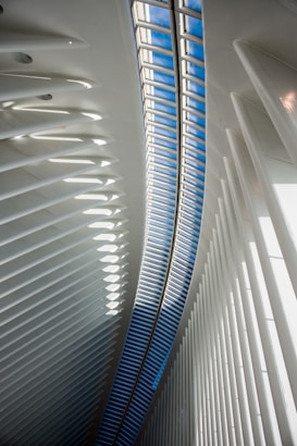 An interior architectural scene featuring a curved ceiling with a long skylight allowing natural light to illuminate the space. The structure is composed of repetitive, vertical white beams which create a sense of depth and dynamism. The skylight reveals a patch of blue sky, contrasting with the white interior.