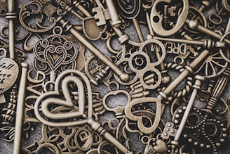 A collection of vintage-style, ornate metal keys with intricate designs, including heart and wing motifs, are scattered across a textured surface. Some keys have unique cutouts and words engraved on them.