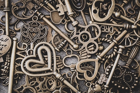 A collection of vintage-style, ornate metal keys with intricate designs, including heart and wing motifs, are scattered across a textured surface. Some keys have unique cutouts and words engraved on them.