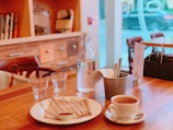 A cozy café setting with a wooden table featuring a plate of crepes drizzled with chocolate sauce and a small dish of syrup. A cup of coffee sits nearby, along with a metal bucket holding utensils and napkins. Two glass cups and a glass bottle of water are also on the table. The background shows a blurred view of a window and soft-focus shelves with books and jars.
