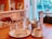 A cozy café setting with a wooden table featuring a plate of crepes drizzled with chocolate sauce and a small dish of syrup. A cup of coffee sits nearby, along with a metal bucket holding utensils and napkins. Two glass cups and a glass bottle of water are also on the table. The background shows a blurred view of a window and soft-focus shelves with books and jars.