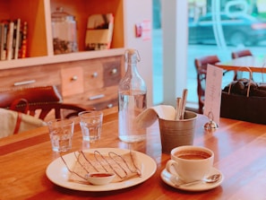 A cozy café setting with a wooden table featuring a plate of crepes drizzled with chocolate sauce and a small dish of syrup. A cup of coffee sits nearby, along with a metal bucket holding utensils and napkins. Two glass cups and a glass bottle of water are also on the table. The background shows a blurred view of a window and soft-focus shelves with books and jars.