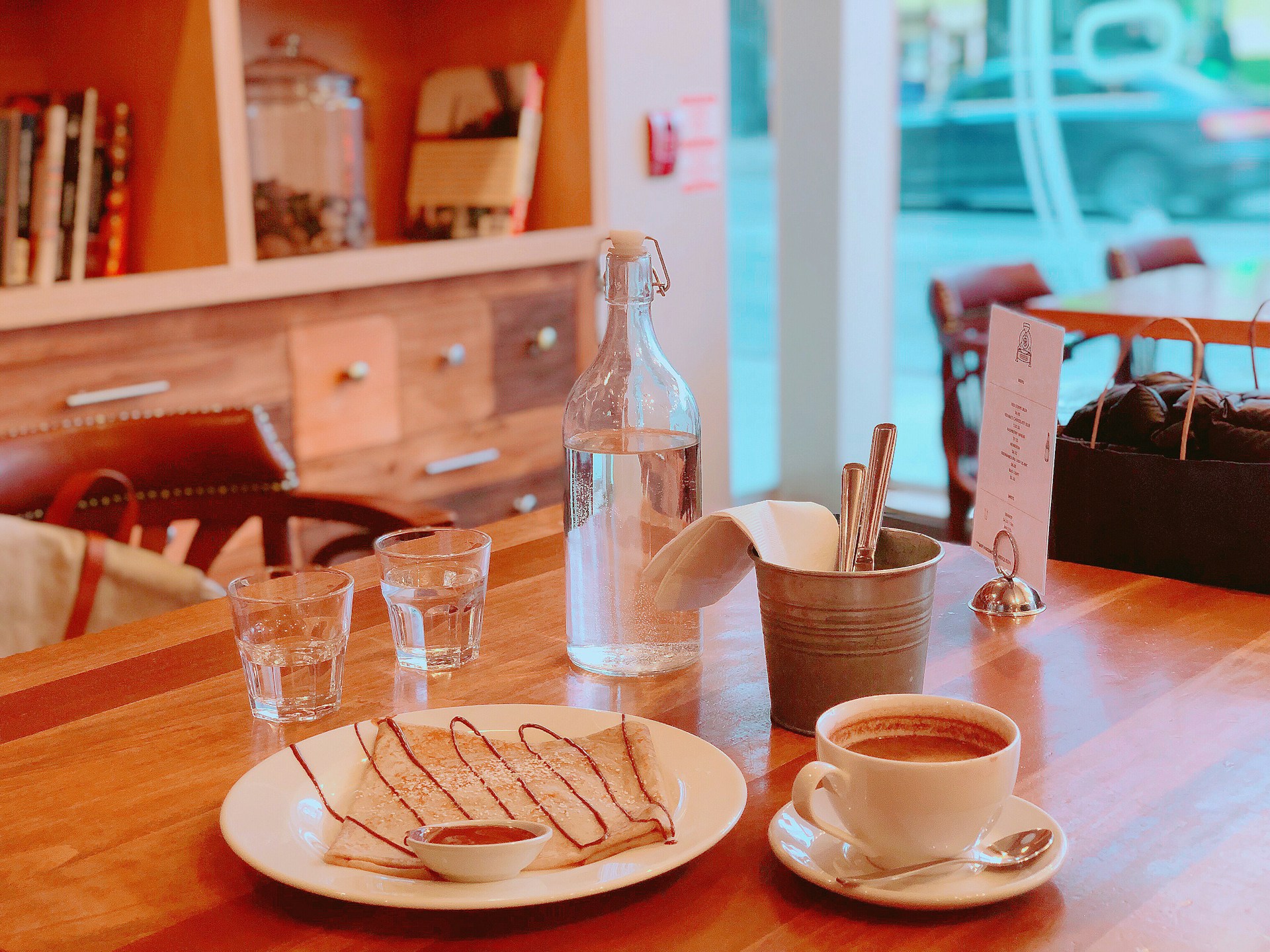 A cozy café setting with a wooden table featuring a plate of crepes drizzled with chocolate sauce and a small dish of syrup. A cup of coffee sits nearby, along with a metal bucket holding utensils and napkins. Two glass cups and a glass bottle of water are also on the table. The background shows a blurred view of a window and soft-focus shelves with books and jars.