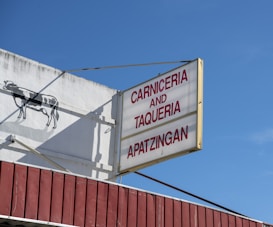 A building sign reads 'Carniceria and Taqueria Apatzingan' in red capital letters on a white background. Adjacent to the sign, a silhouette of a cow is painted on the white wall. Below, there is a maroon-colored awning. The sky is clear and blue.