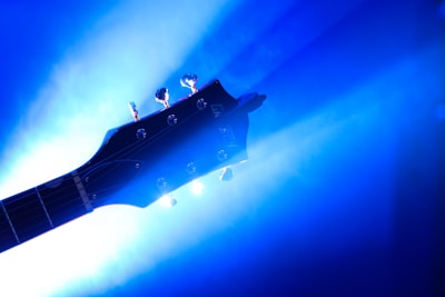 A silhouette of an electric guitar headstock is set against a vibrant blue background with dramatic lighting. The light reflects off the tuning pegs, creating an ethereal and artistic effect.