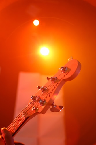 A close-up of a guitar headstock with tuning pegs is illuminated by a warm, orange stage light in the background. The brand name is visible on the headstock, and spotlights create a vibrant and energetic atmosphere.