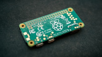 A Raspberry Pi Zero W circuit board is seen on a dark surface. It is green with various gold and white printed details, including the Raspberry Pi logo. The board contains numerous gold connectors and ports, as well as white printed text and symbols.