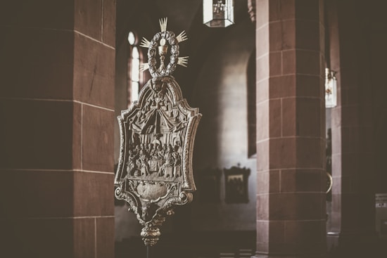 An ornate religious artifact, featuring detailed carvings and symbols, is prominently displayed in a dimly lit interior of a church. The background includes large stone columns and softly filtered natural light coming through an arched window.