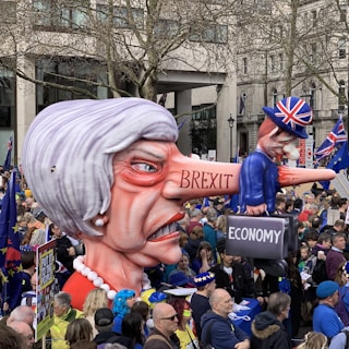 A large caricature of a woman with exaggerated features, including a long nose, labeled 'BREXIT' is being held by a crowd in a protest setting. On the nose is a smaller figure wearing a Union Jack hat, holding a suitcase labeled 'ECONOMY'. The crowd is diverse and holds flags and signs, set in an urban environment with trees and buildings visible in the background.