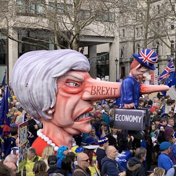 A large caricature of a woman with exaggerated features, including a long nose, labeled 'BREXIT' is being held by a crowd in a protest setting. On the nose is a smaller figure wearing a Union Jack hat, holding a suitcase labeled 'ECONOMY'. The crowd is diverse and holds flags and signs, set in an urban environment with trees and buildings visible in the background.