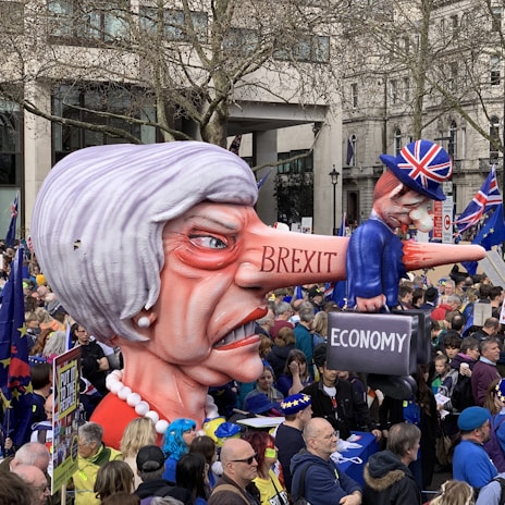 A large caricature of a woman with exaggerated features, including a long nose, labeled 'BREXIT' is being held by a crowd in a protest setting. On the nose is a smaller figure wearing a Union Jack hat, holding a suitcase labeled 'ECONOMY'. The crowd is diverse and holds flags and signs, set in an urban environment with trees and buildings visible in the background.