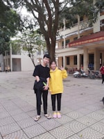 Two people are standing together outside, with one wearing a black outfit and the other a yellow sweater. They are smiling and making peace signs. The background shows a large tree, a tiled courtyard, and a beige building with multiple floors and a red banner. There are also a few motorbikes parked nearby.