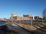 A large, industrial complex featuring red brick buildings and tall chimneys is visible. The foreground is a landscaped area with neatly arranged rows of wooden posts and soil. A blue sky provides a clear backdrop, adding a sense of openness to the scene. Sparse trees line the right edge of the image.