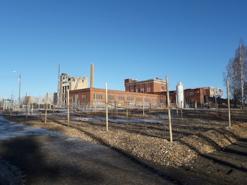 A large, industrial complex featuring red brick buildings and tall chimneys is visible. The foreground is a landscaped area with neatly arranged rows of wooden posts and soil. A blue sky provides a clear backdrop, adding a sense of openness to the scene. Sparse trees line the right edge of the image.