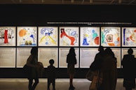Silhouettes of people observing colorful and abstract artwork arranged in a gallery display. The artwork features various bold colors and dynamic shapes, with text panels below each visual piece.