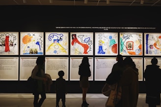 Silhouettes of people observing colorful and abstract artwork arranged in a gallery display. The artwork features various bold colors and dynamic shapes, with text panels below each visual piece.