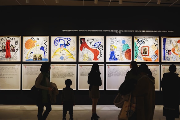 Silhouettes of people observing colorful and abstract artwork arranged in a gallery display. The artwork features various bold colors and dynamic shapes, with text panels below each visual piece.