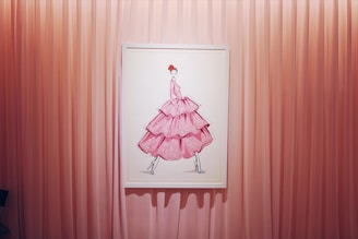 A framed artwork of a fashion illustration depicting a figure in a voluminous, layered pink dress with polka dots. The background consists of vertical pink curtains that create a soft and elegant setting.