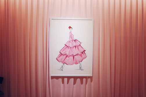 A framed artwork of a fashion illustration depicting a figure in a voluminous, layered pink dress with polka dots. The background consists of vertical pink curtains that create a soft and elegant setting.