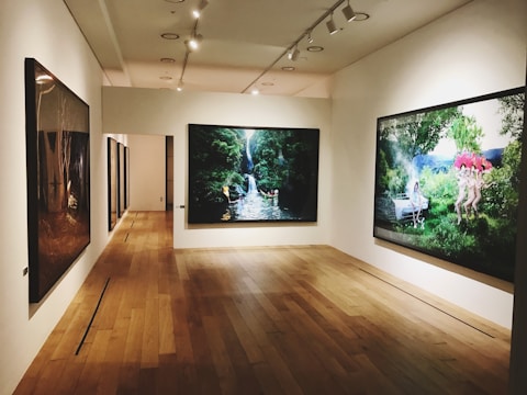 A gallery room with wooden flooring and recessed lighting displays large framed artworks on the walls. The central piece features a lush green landscape with a waterfall, while another artwork shows figures in a natural setting among trees and flowers.