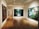 A gallery room with wooden flooring and recessed lighting displays large framed artworks on the walls. The central piece features a lush green landscape with a waterfall, while another artwork shows figures in a natural setting among trees and flowers.