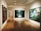 A gallery room with wooden flooring and recessed lighting displays large framed artworks on the walls. The central piece features a lush green landscape with a waterfall, while another artwork shows figures in a natural setting among trees and flowers.