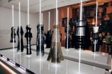 A display of elegant dresses with metallic and intricate patterns, showcased under vertical light beams in a modern exhibition setting. The background features a collage of photographs and a diverse group of people observing the installation.