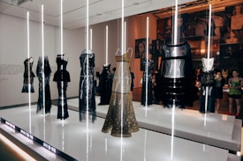 A display of elegant dresses with metallic and intricate patterns, showcased under vertical light beams in a modern exhibition setting. The background features a collage of photographs and a diverse group of people observing the installation.