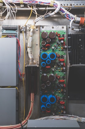 A detailed view of an electrical or electronic circuit board, complete with various components such as capacitors, resistors, and wires. The wires are neatly arranged and secured at the top of the panel, leading to connections on the circuit board. The board has a noticeable number of red and blue components, likely indicating different types of electronic elements.