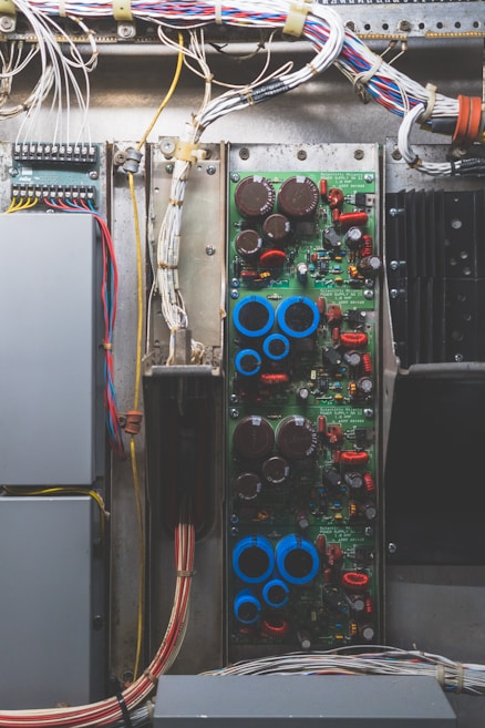 A detailed view of an electrical or electronic circuit board, complete with various components such as capacitors, resistors, and wires. The wires are neatly arranged and secured at the top of the panel, leading to connections on the circuit board. The board has a noticeable number of red and blue components, likely indicating different types of electronic elements.