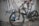 A mountain bike is positioned against a wall with graffiti art. The bike features thick tires, full suspension, and a sleek design. The graffiti includes stylized text and abstract figures, adding an urban, edgy feel to the scene. The ground is littered with debris, enhancing the raw atmosphere.