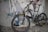 A mountain bike is positioned against a wall with graffiti art. The bike features thick tires, full suspension, and a sleek design. The graffiti includes stylized text and abstract figures, adding an urban, edgy feel to the scene. The ground is littered with debris, enhancing the raw atmosphere.