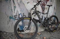 A mountain bike is positioned against a wall with graffiti art. The bike features thick tires, full suspension, and a sleek design. The graffiti includes stylized text and abstract figures, adding an urban, edgy feel to the scene. The ground is littered with debris, enhancing the raw atmosphere.