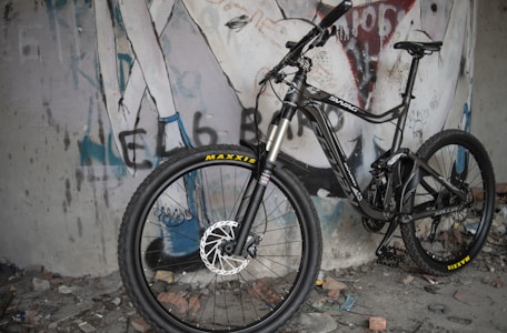 A mountain bike is positioned against a wall with graffiti art. The bike features thick tires, full suspension, and a sleek design. The graffiti includes stylized text and abstract figures, adding an urban, edgy feel to the scene. The ground is littered with debris, enhancing the raw atmosphere.
