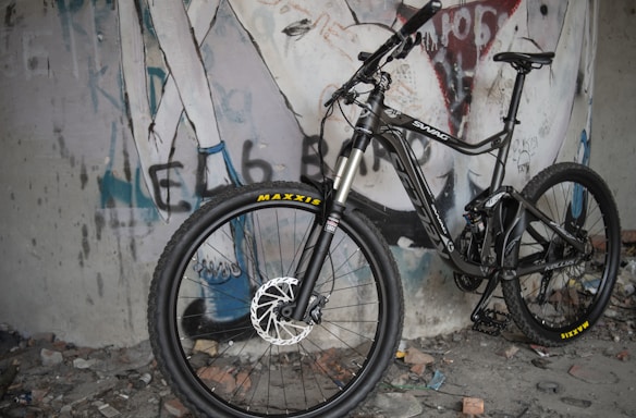 A mountain bike is positioned against a wall with graffiti art. The bike features thick tires, full suspension, and a sleek design. The graffiti includes stylized text and abstract figures, adding an urban, edgy feel to the scene. The ground is littered with debris, enhancing the raw atmosphere.