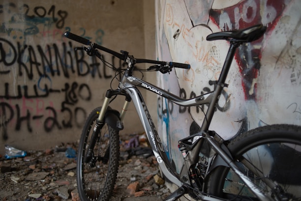 A mountain bike with a black and silver frame is propped against a wall covered in graffiti. The area appears rugged with scattered debris and rocks on the ground. The graffiti features various words and symbols, including large, bold text.