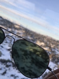 A pair of aviator sunglasses is held up against a snowy landscape. The lenses reflect parts of the light sky and blurred snow-covered terrain in the background, creating a contrast between the greenish tint of the glasses and the white snow.