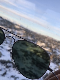 A pair of aviator sunglasses is held up against a snowy landscape. The lenses reflect parts of the light sky and blurred snow-covered terrain in the background, creating a contrast between the greenish tint of the glasses and the white snow.