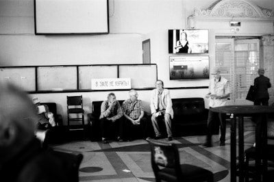 A black and white image depicts a waiting area with several people sitting on black couches. There is a sign with Russian text, two television screens, and ornate architectural details along the walls and ceiling. The floor is patterned, and people are casually dressed, engaging in conversation or reading.