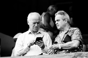 Two elderly men are standing close together in conversation. One of them is holding and looking at a smartphone, while the other appears to be explaining or gesturing towards something. They are engaged and seem focused on the discussion. Behind them, other people can be seen in blurred background.
