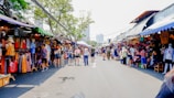 A busy outdoor market scene with numerous stalls displaying colorful clothing and merchandise. People walk along the pathway, engaging with vendors and browsing items. The market is set under bright sunlight with trees providing partial shade. The atmosphere is lively with a mix of tourists and locals.