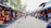 A busy outdoor market scene with numerous stalls displaying colorful clothing and merchandise. People walk along the pathway, engaging with vendors and browsing items. The market is set under bright sunlight with trees providing partial shade. The atmosphere is lively with a mix of tourists and locals.