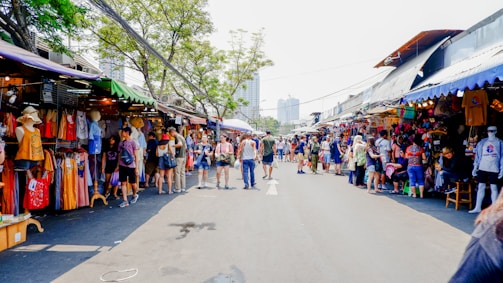 A busy outdoor market scene with numerous stalls displaying colorful clothing and merchandise. People walk along the pathway, engaging with vendors and browsing items. The market is set under bright sunlight with trees providing partial shade. The atmosphere is lively with a mix of tourists and locals.