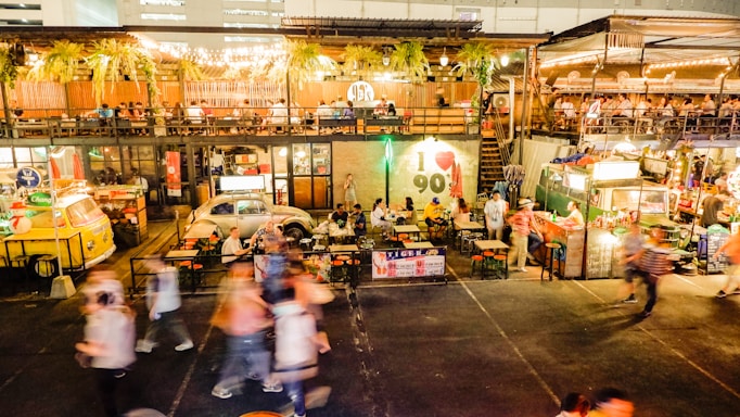 An outdoor food market is bustling with people walking around and others sitting at tables enjoying meals. The scene includes vibrant food stalls, colorful lighting, and greenery hanging from above. The environment looks lively and social, with a mix of retro and modern decor.